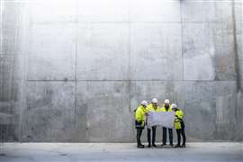 A group of four engineers in hard hats and yellow hi-vis jackets examine plans against the backdrop of a concrete wall