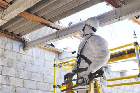 A worker in full protective clothing and respiratory protection equipment stands on a stepladder to look at asbestos-containing roof sheets.