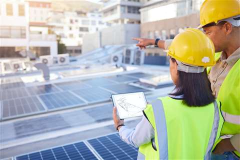 A female and male engineer on the rooftop of a new building with a roof covered in solar panels inspect plans on a digital device